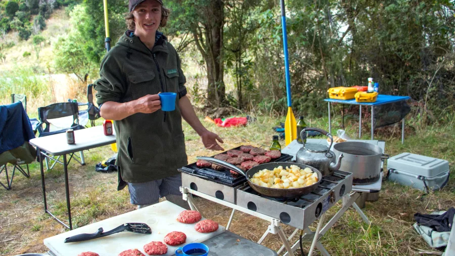 Outdoor chef preparing burgers and potatoes at riverside camp kitchen
