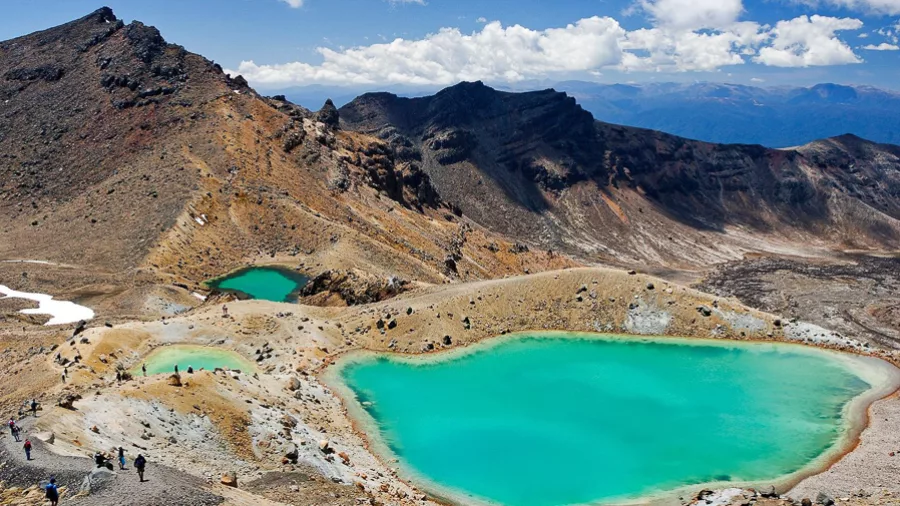 Hikers trek beside the Emerald Lakes on the Tongariro Alpine Crossing, surrounded by volcanic mountains and dramatic alpine scenery in New Zealand.