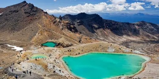 Hikers trek beside the Emerald Lakes on the Tongariro Alpine Crossing, surrounded by volcanic mountains and dramatic alpine scenery in New Zealand.