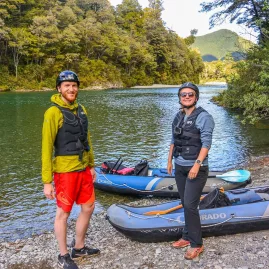 Two kayakers smiling beside inflatable rafts on the edge of the Pelorus River