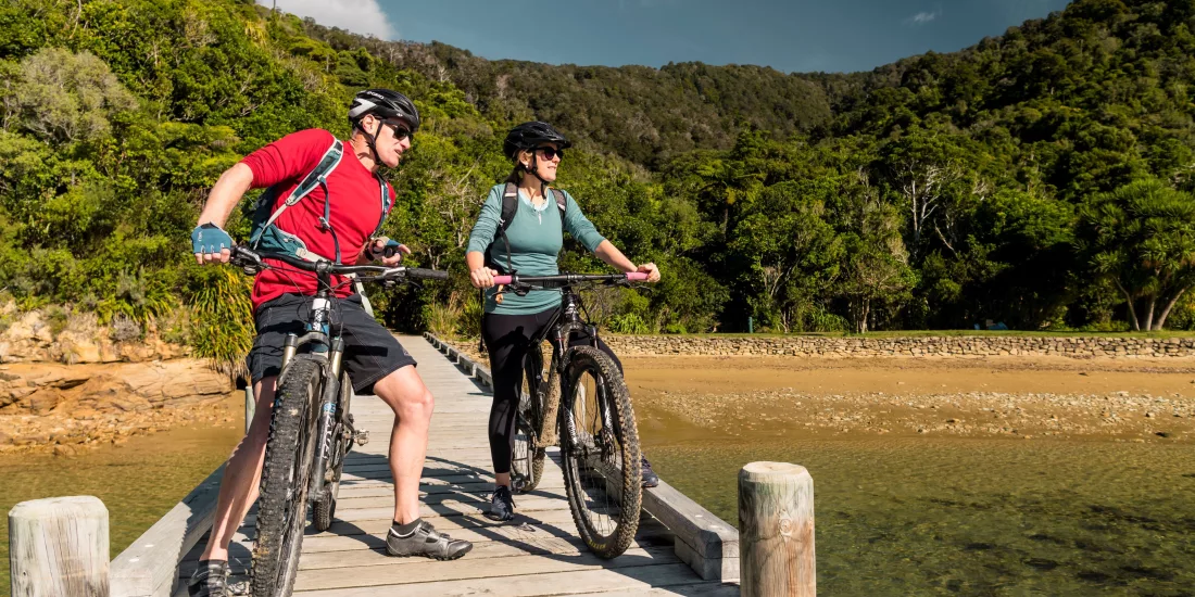 Couple with mountain bikes pausing at the edge of the Ship Cove jetty with forest in the background.