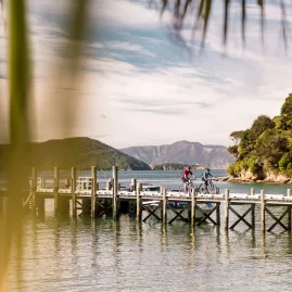 Cyclists riding on the jetty at Ship Cove, Queen Charlotte Track, with Marlborough Sounds in the background.