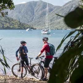 Two cyclists resting near the beach at Furneaux Lodge with yachts anchored in the Marlborough Sounds.