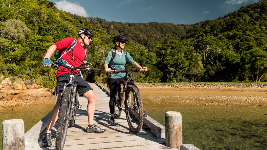 Couple with mountain bikes pausing at the edge of the Ship Cove jetty with forest in the background.