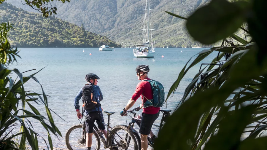Two cyclists resting near the beach at Furneaux Lodge with yachts anchored in the Marlborough Sounds.