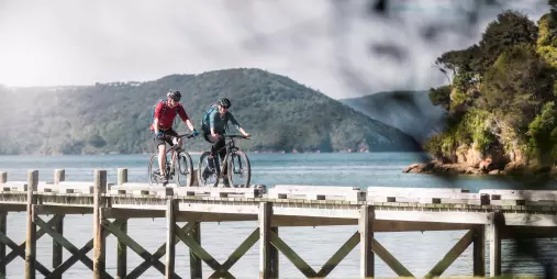 Two cyclists riding along the pier at Ship Cove, framed by forested hills and calm waters.