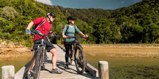 Couple with mountain bikes pausing at the edge of the Ship Cove jetty with forest in the background.