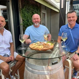 Three men enjoying a shared platter and white wine at a wine barrel table