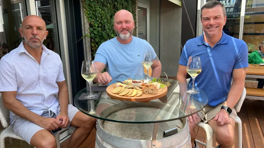 Three men enjoying a shared platter and white wine at a wine barrel table