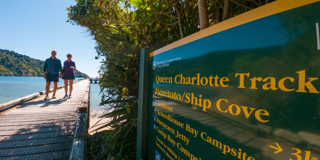 Couple walking past Queen Charlotte Track sign near the water at Ship Cove