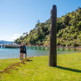 Visitors standing near a carved wooden post by the wharf at Ship Cove
