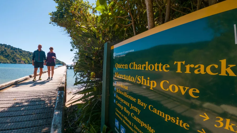 Couple walking past Queen Charlotte Track sign near the water at Ship Cove