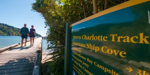 Couple walking past Queen Charlotte Track sign near the water at Ship Cove