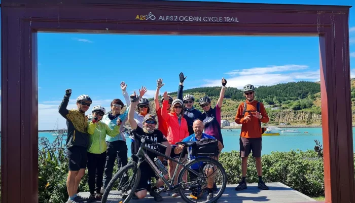 Group of cyclists celebrating at the Alps 2 Ocean Cycle Trail finish line in Oamaru, New Zealand, with bikes and raised hands.