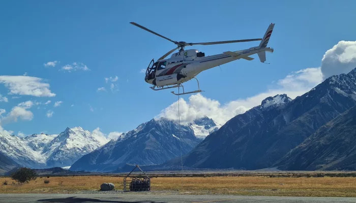 Helicopter transporting cargo with a sling load in the Tasman River valley, surrounded by snow-capped mountains.