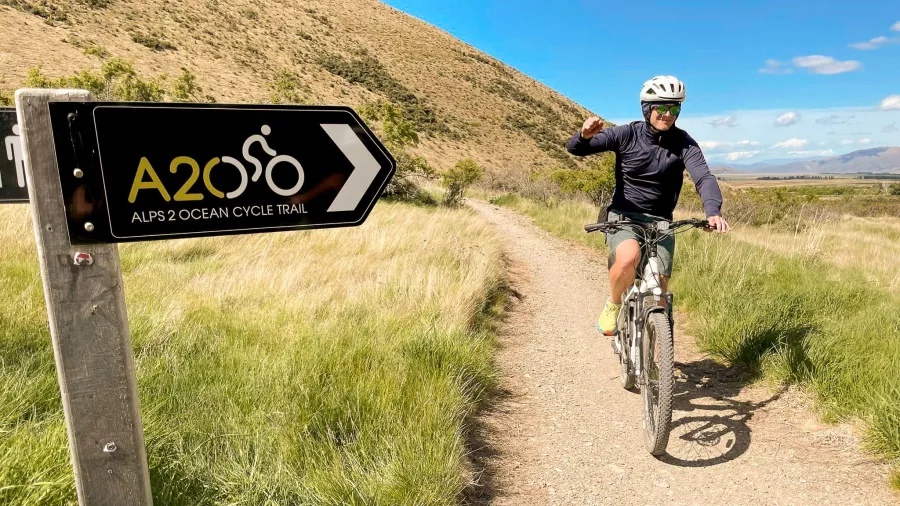 Cyclist riding past an Alps 2 Ocean Cycle Trail sign on a gravel path through grassy hills in New Zealand.