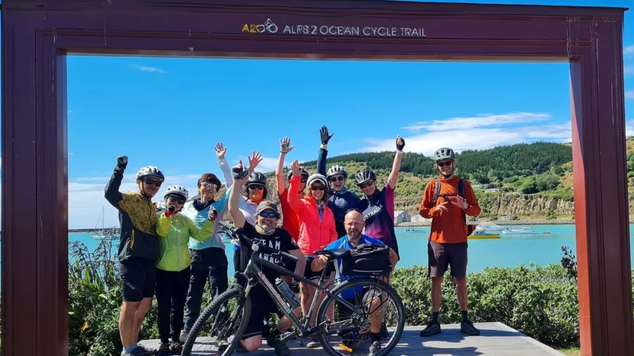 Group of cyclists celebrating at the Alps 2 Ocean Cycle Trail finish line in Oamaru, New Zealand, with bikes and raised hands.