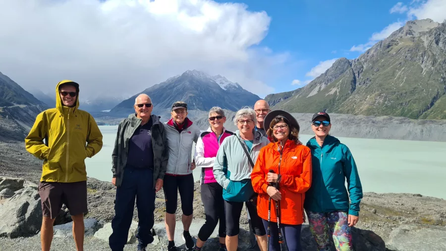 Smiling group of hikers posing at the Tasman Glacier viewpoint with glacial lake and mountains behind.