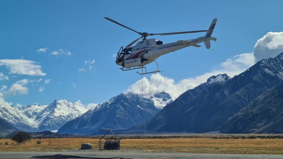 Helicopter transporting cargo with a sling load in the Tasman River valley, surrounded by snow-capped mountains.