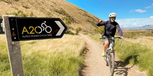 Cyclist riding past an Alps 2 Ocean Cycle Trail sign on a gravel path through grassy hills in New Zealand.