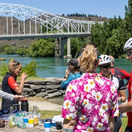Cyclists enjoying a refreshment break beside a river with a bridge in the background.
