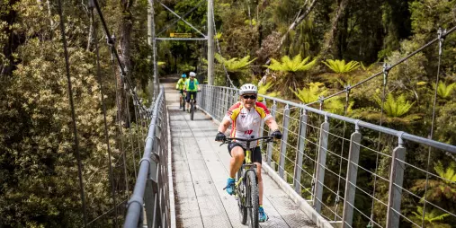 Cyclists crossing a suspension bridge through rainforest on the West Coast of New Zealand.
