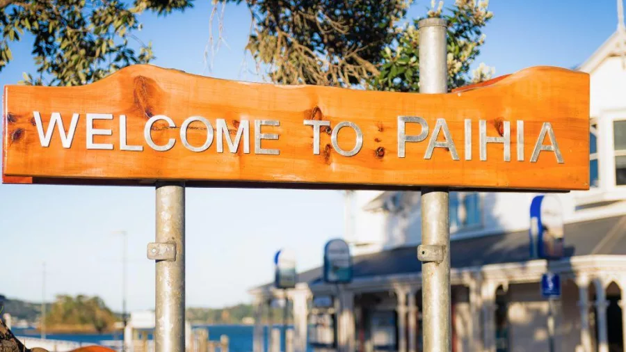 Wooden welcome sign for Paihia with shops and waterfront in the background