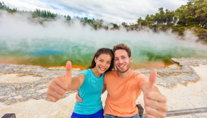 Smiling couple giving thumbs up in front of the colourful Champagne Pool at Wai-O-Tapu Thermal Wonderland near Rotorua