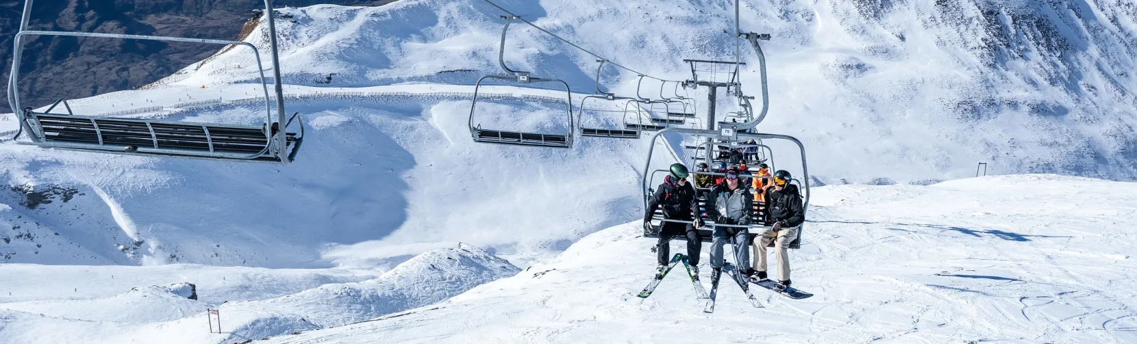 Skiers on chairlift with sweeping alpine views at Treble Cone