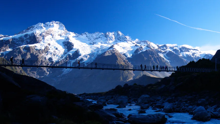 People crossing a swing bridge at sunset with Aoraki Mount Cook in the background