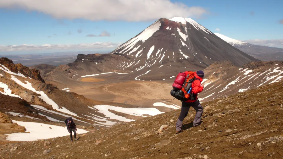 Hiker climbs Mount Tongariro with snow patches and Mt Ngauruhoe towering in the background in Tongariro National Park, New Zealand.
