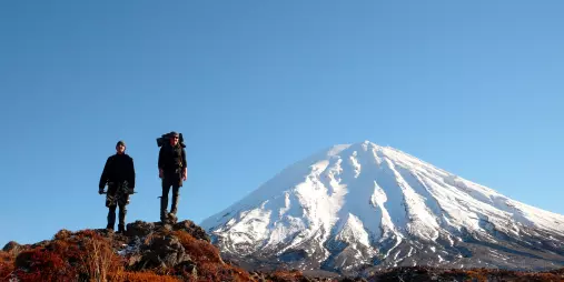 Two hikers stand near Mount Ngauruhoe, snow-covered and under a clear blue sky in Tongariro National Park, New Zealand.
