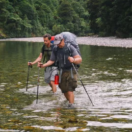 Two hikers crossing a shallow river en route to the Ballroom Overhang in Paparoa National Park