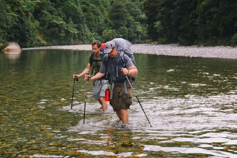 Two hikers crossing a shallow river en route to the Ballroom Overhang in Paparoa National Park