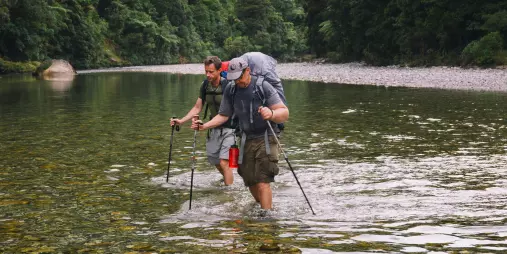 Two hikers crossing a shallow river en route to the Ballroom Overhang in Paparoa National Park