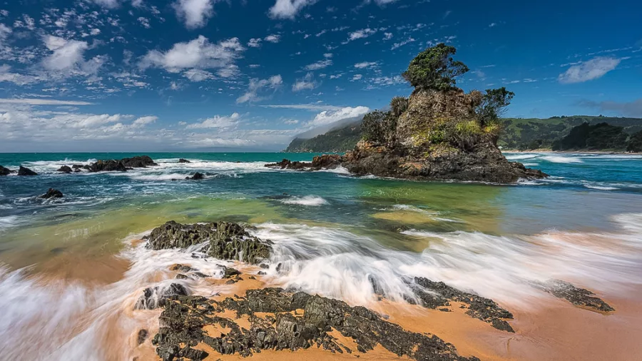 Rocky islet surrounded by turquoise surf on Coromandel coast under blue sky