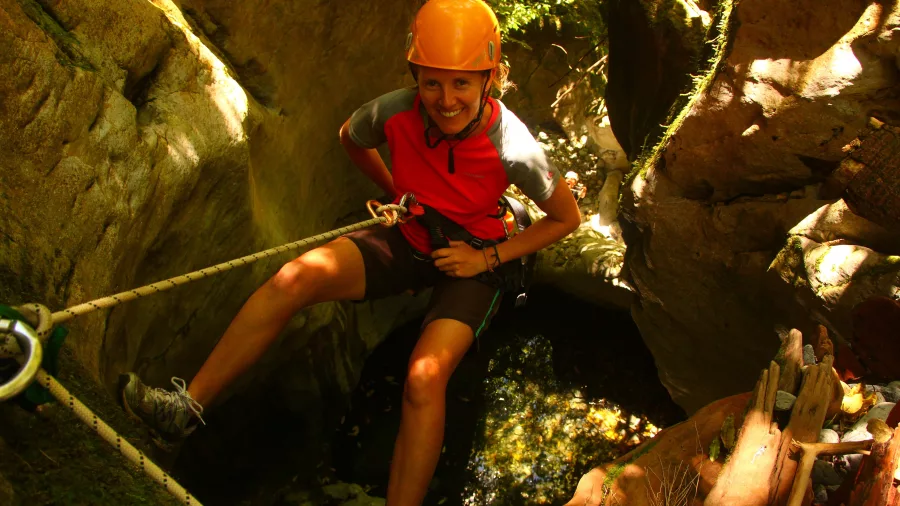 Smiling woman in orange helmet abseiling above a rocky pool in Blue Creek canyon