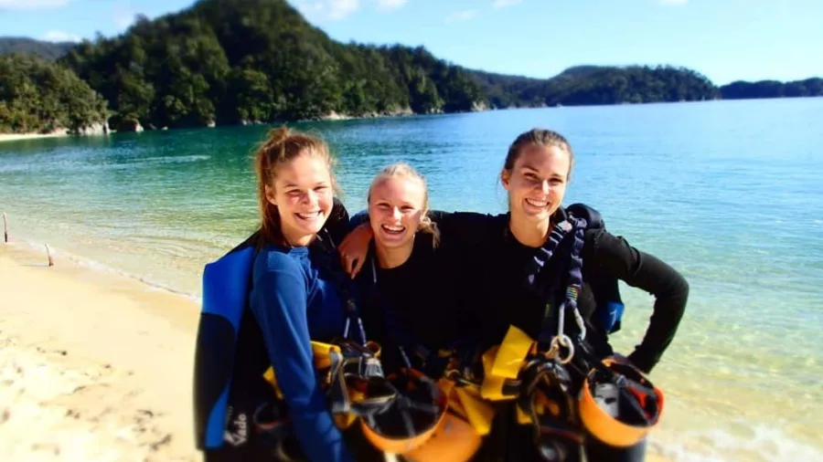 Three smiling canyoners on golden sand beach in Abel Tasman with wetsuits and helmets