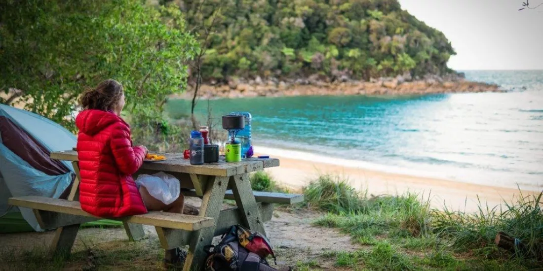 Camper in red jacket enjoying a peaceful beachfront breakfast in Abel Tasman