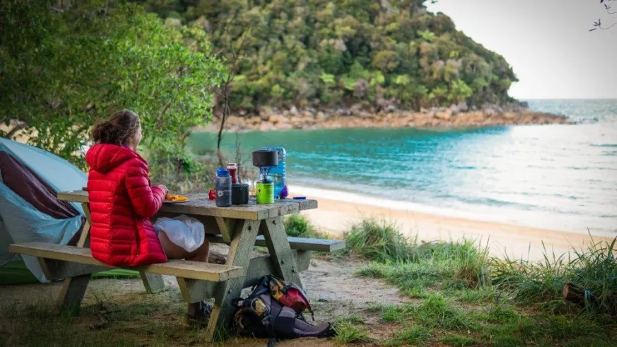 Camper in red jacket enjoying a peaceful beachfront breakfast in Abel Tasman