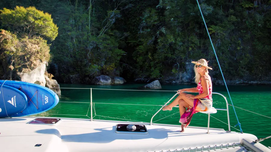 Woman in sunhat relaxing on the front deck of catamaran near bush-clad cove