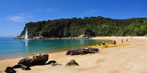 Kayaks pulled up on golden sand beach in Abel Tasman National Park with people preparing for a sailing trip