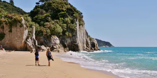 Two people walking along Awaroa Beach with cliffs and native bush in Abel Tasman National Park