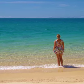 Woman in a floral dress standing on a beach in Abel Tasman, looking out at the turquoise ocean