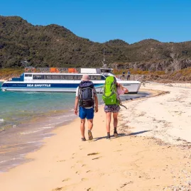 Two hikers walking along the beach toward an Abel Tasman Sea Shuttle boat during the Discoverer Day tour