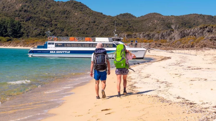 Two hikers walking along the beach toward an Abel Tasman Sea Shuttle boat during the Discoverer Day tour