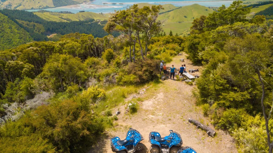 Quad bikes and a Polaris parked at a picnic lookout on the Bayview Circuit, with riders gathered around a bench enjoying coastal views in Cable Bay, New Zealand.