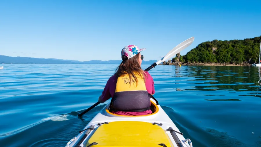 Woman paddling solo kayak toward lush green coastline in Golden Bay