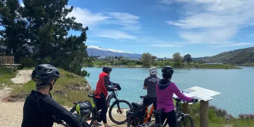 Group of cyclists stopping to read an information sign beside a lake in Cromwell with mountain views