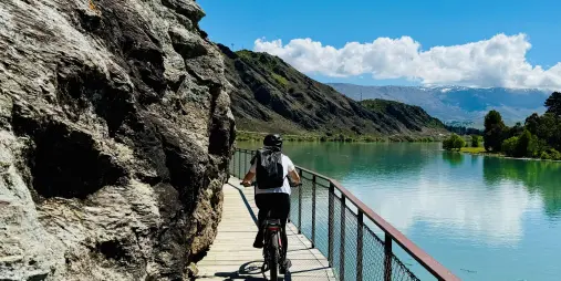 Cyclist riding cliffside path above Lake Dunstan with mountain backdrop